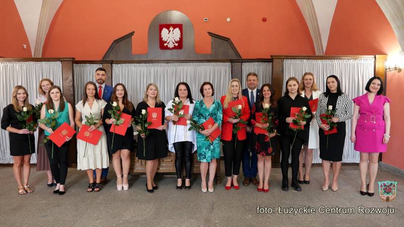 Czternaście kobiet i dwóch mężczyzn w odświętnych strojach pozuje do zdjęcia. Większość trzyma w rękach białe róże oraz czerwone teczki z herbem miasta. Nad nimi widać godło narodowe Polski.